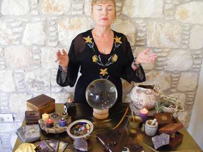 A person with their face obscured is standing behind a table filled with various objects including a crystal ball, boxes, and candles against a stone wall background.