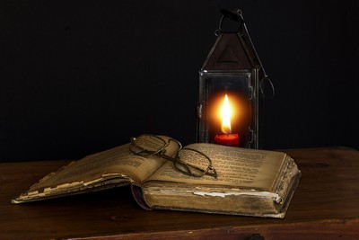 An open, worn book rests on a wooden surface with a pair of round spectacles placed on top of the right page. To the right of the book stands a metal-framed lantern with glass panels, inside which a single candle burns brightly, illuminating the scene with a warm glow against a dark background. The setting evokes a sense of antiquity, magic and the pursuit of magical knowledge.