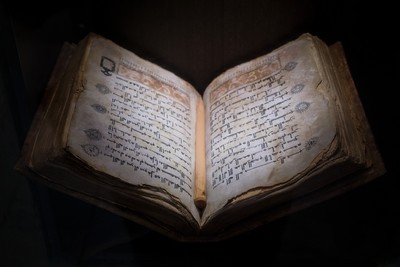 An open ancient Grimoire manuscript is displayed, resting on a dark surface with its pages slightly worn and yellowed, indicating age. The manuscript contains spells, notations and symbols consistent with a Witches Book of Shadows. The lighting casts shadows across the pages, highlighting the texture of the paper and the handwritten content.