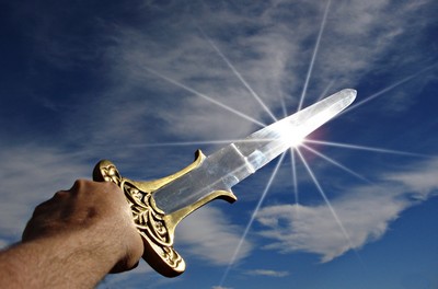 A person’s hand holding a transparent sword with a golden hilt up towards the sky. The sun shines brightly behind the blade, creating a lens flare effect. The background is a clear blue sky with wispy white clouds and contrails from an airplane.
