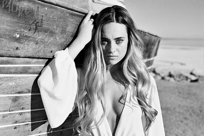 A black and white photo of a person with long hair standing in front of a wooden structure, with their face obscured by a blurred rectangular area, set against a backdrop of a calm beach.