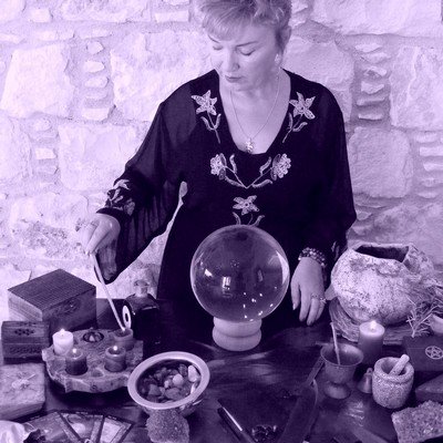 A black and white photo of a person sitting at a table with various objects, including a crystal ball, candles, and a bowl of stones.